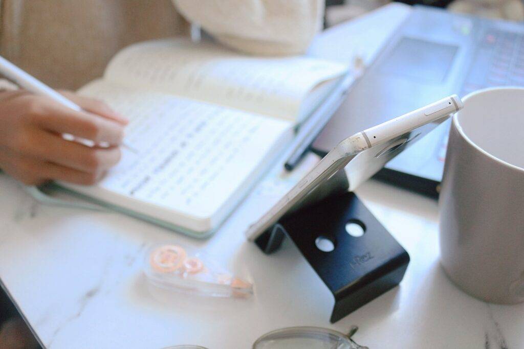 Hand writing notes in a notebook beside a laptop and phone