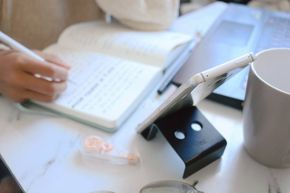 Hand writing notes in a notebook beside a laptop and phone